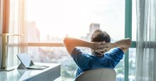 Man is sitting contentedly looking outside from a tall office building with his arms behind his head, leaning back and has a laptop on a desk