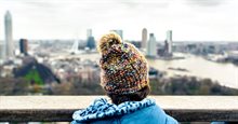 Child wearing a hat and a warm blue coat stands on top of a tall building and looks out over a city